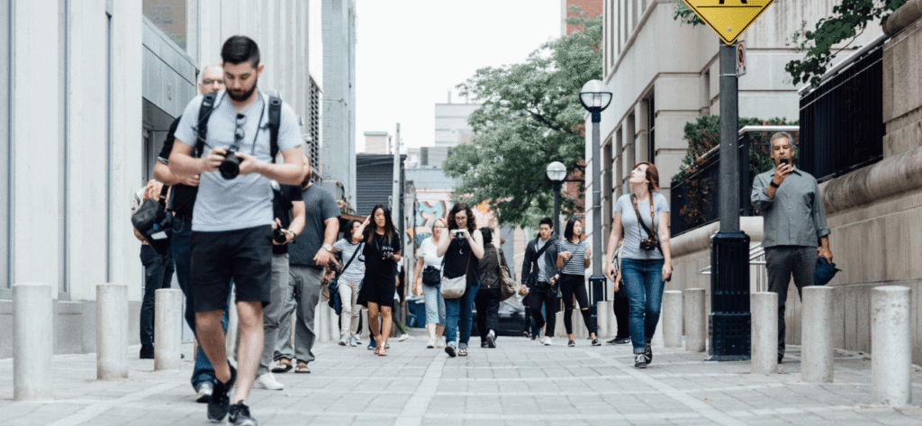 Pedestrians carrying cameras and smartphones walk along a busy urban sidewalk between city buildings.