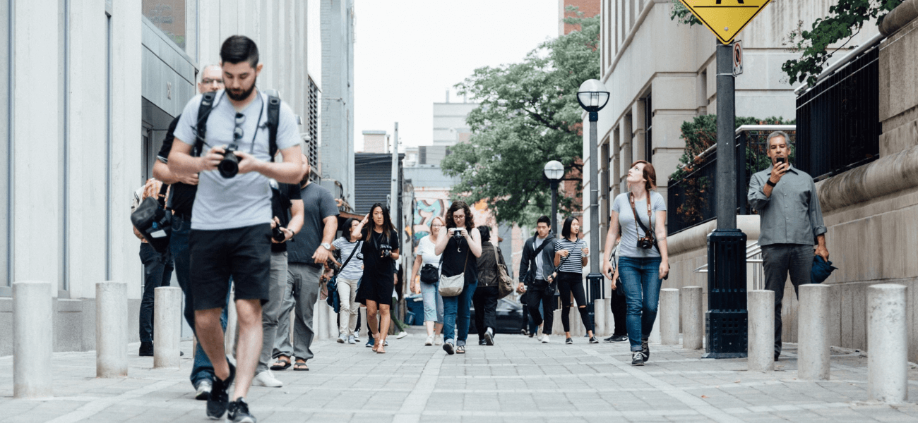 Pedestrians carrying cameras and smartphones walk along a busy urban sidewalk between city buildings.