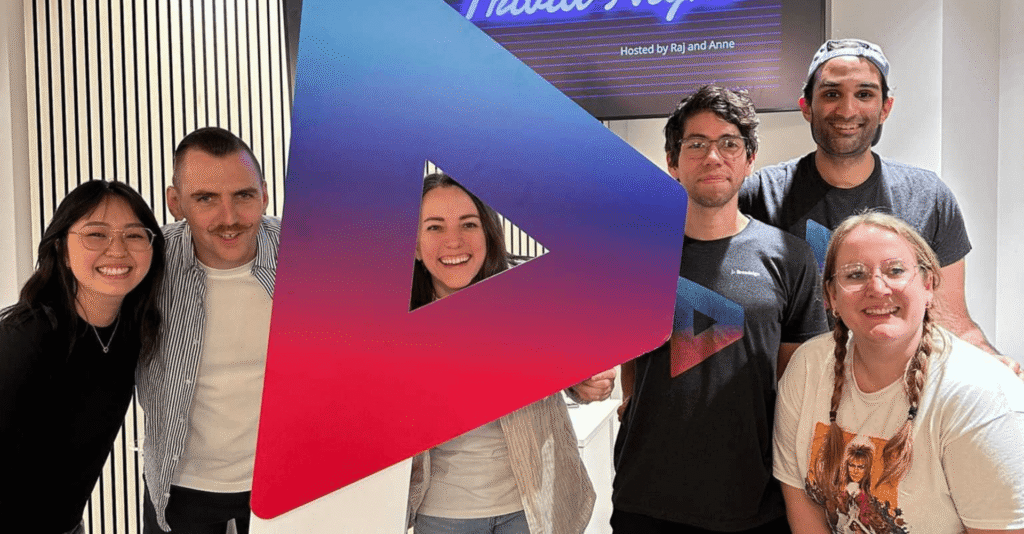 Group of six colleagues smiling together around a large colorful Broadsign logo display in an office setting.