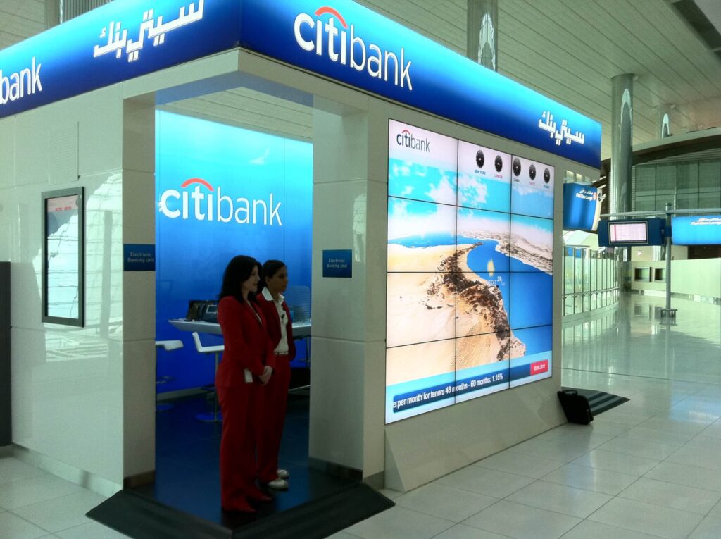 Citibank digital advertising display inside an airport terminal, with two airline staff members standing nearby.