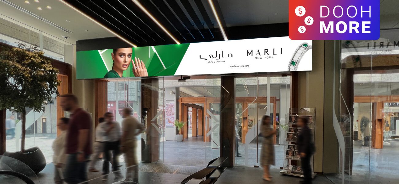 Large digital billboard featuring a MARLI New York jewelry ad above the entrance of a busy shopping center.