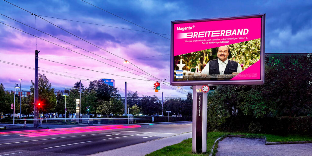 Digital roadside billboard featuring a Magenta broadband advertisement at a city intersection at dusk.