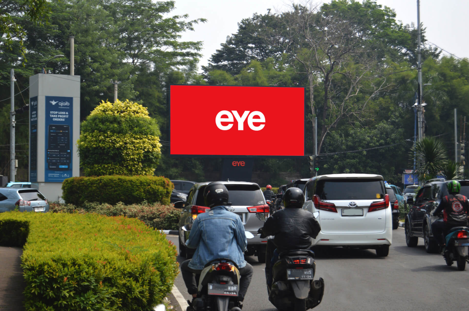 Large Eye digital billboard overlooking cars and motorcyclists on a busy tree-lined road.