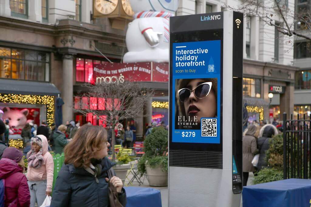 Digital street advertising display in a busy shopping area promotes an interactive holiday storefront for eyewear, featuring a QR code for shoppers to scan and buy.