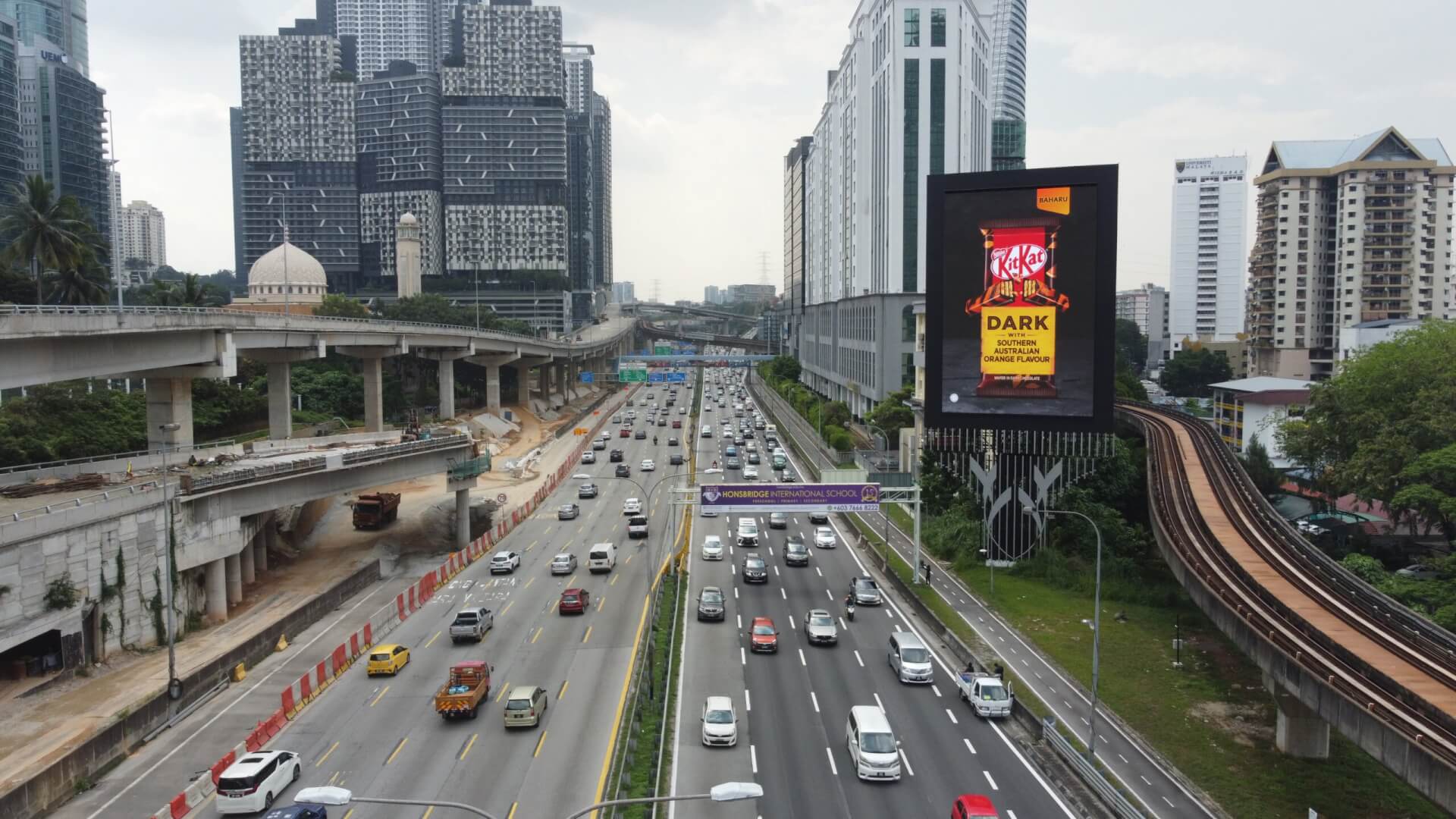 Digital billboard beside a busy multi-lane highway and elevated rail line in a modern city.