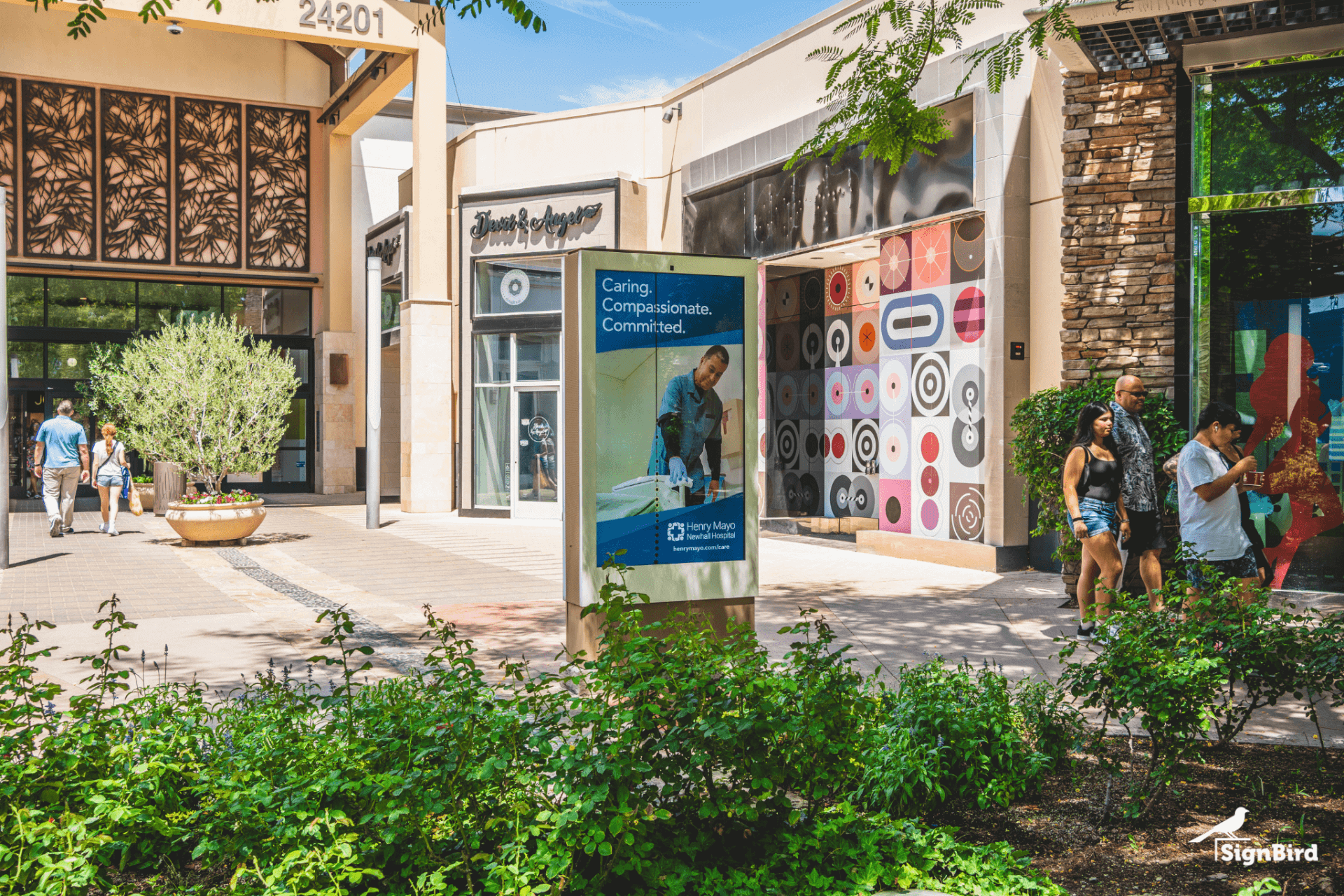 Digital advertising display in an outdoor shopping center, surrounded by storefronts, greenery, and pedestrians.
