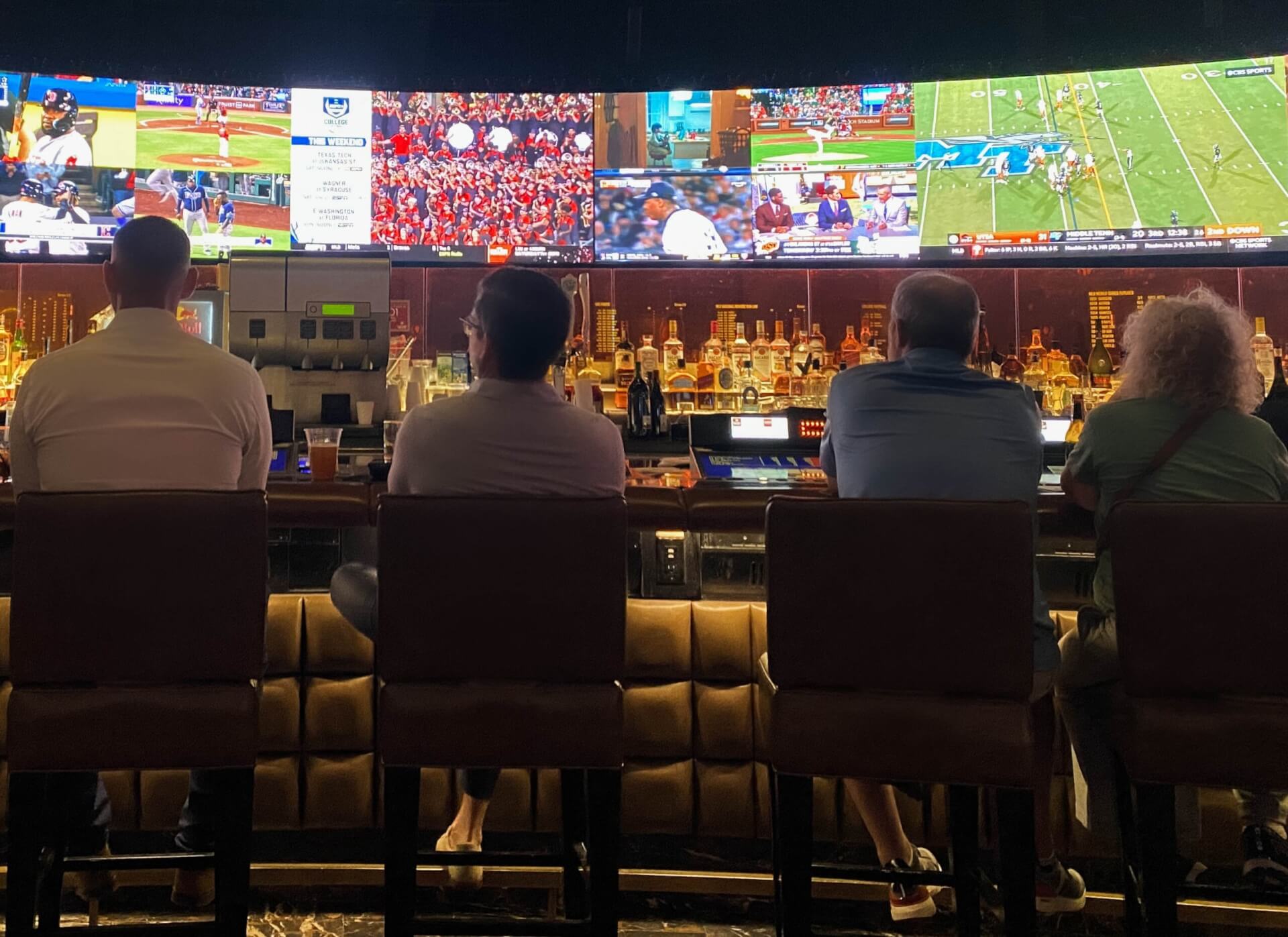 Four people seated at a bar watching live sports on multiple television screens.