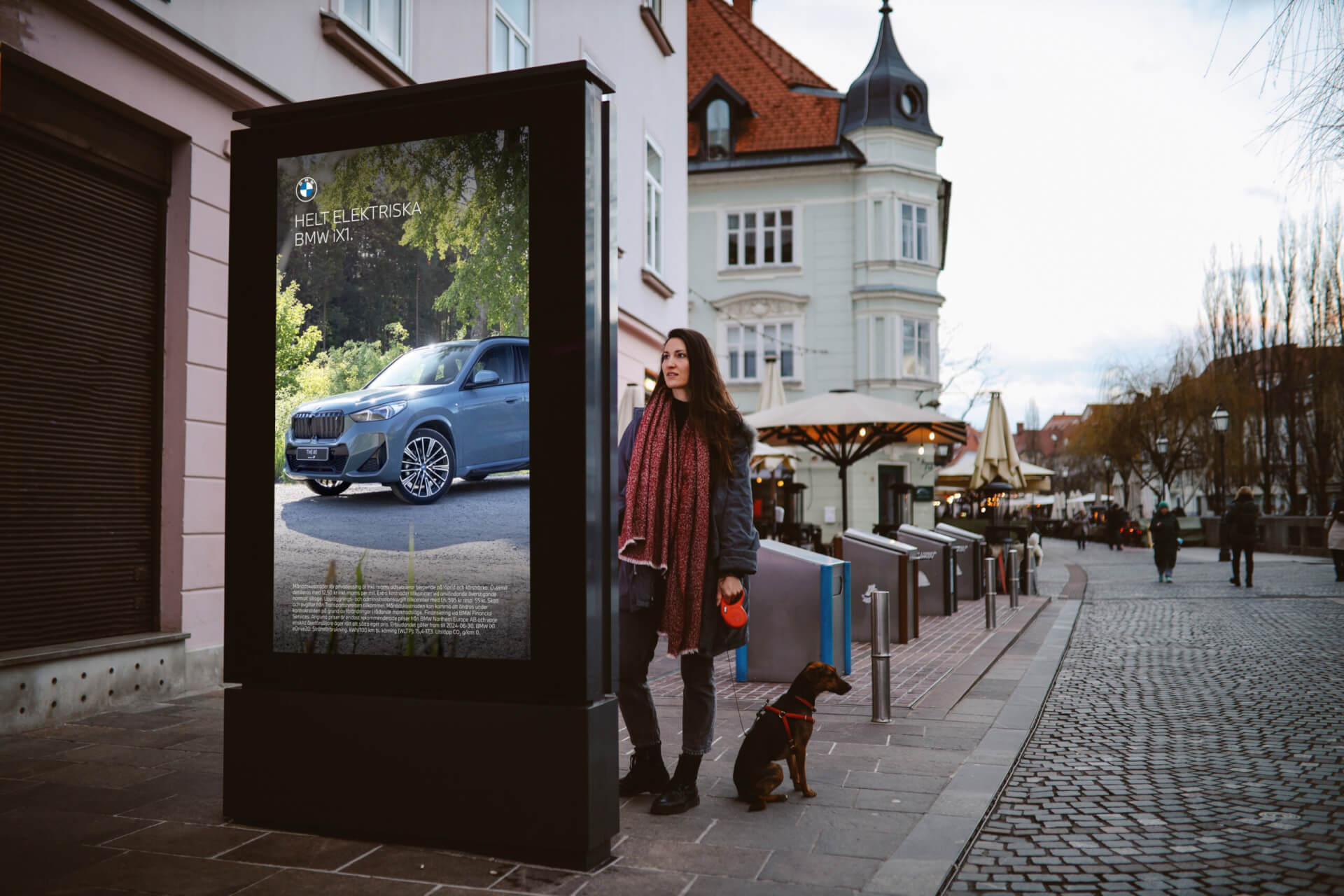 Man walking a small dog past a digital street display featuring a car advertisement on a cobblestone city street.