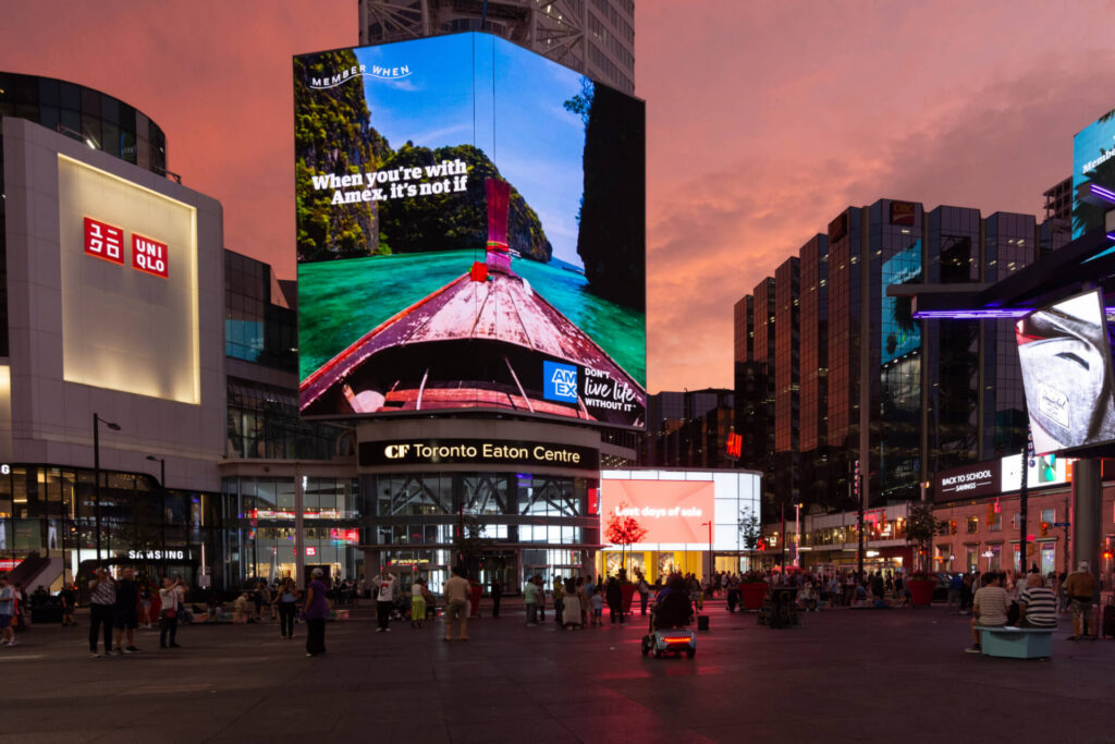 Busy urban plaza surrounded by large digital billboards displaying colorful advertisements above crowds of pedestrians.