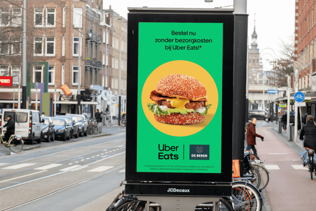 Uber Eats and De Beren digital out-of-home ad featuring a burger and free delivery offer at a city transit shelter.