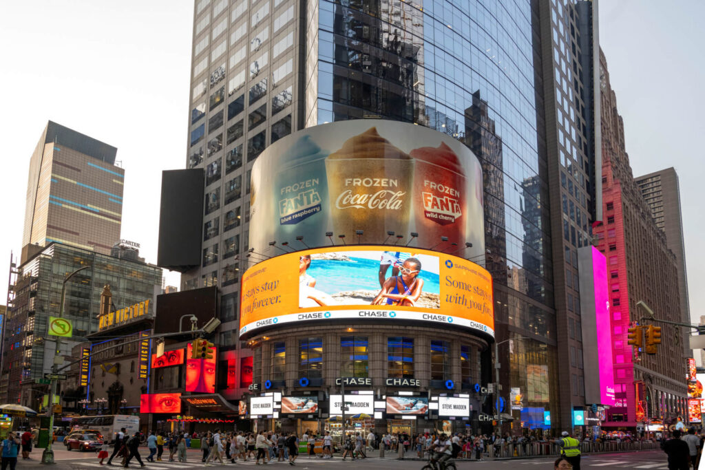 Large digital billboards on a curved building in a busy city intersection display Coca-Cola and Chase advertisements, surrounded by high-rise buildings.