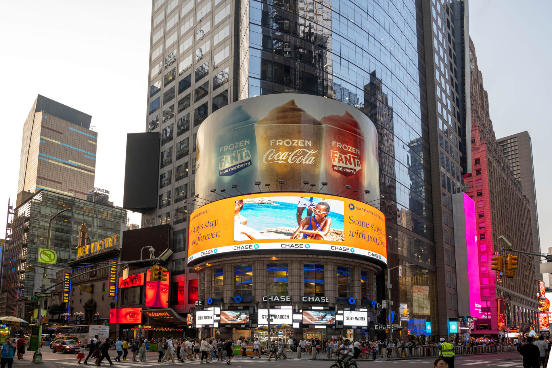 Large digital billboards on a curved building in a busy city intersection display Coca-Cola and Chase advertisements, surrounded by high-rise buildings.