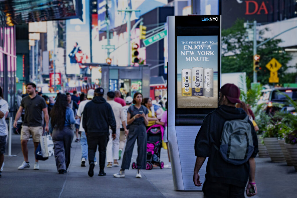 A busy New York City sidewalk with pedestrians walking past a LinkNYC digital kiosk displaying a Corona beer advertisement. The kiosk stands prominently in the foreground against a backdrop of bright city lights, storefronts, and traffic, illustrating digital out-of-home advertising in a high-footfall urban environment.