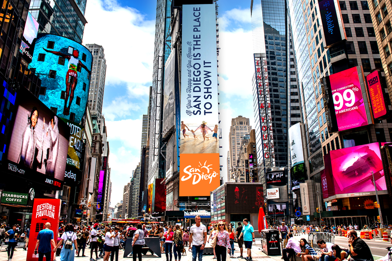 A vibrant Times Square scene featuring a towering San Diego tourism digital billboard with the message “San Diego is the place to show it,” surrounded by colorful advertisements, skyscrapers, pedestrians, and city traffic.