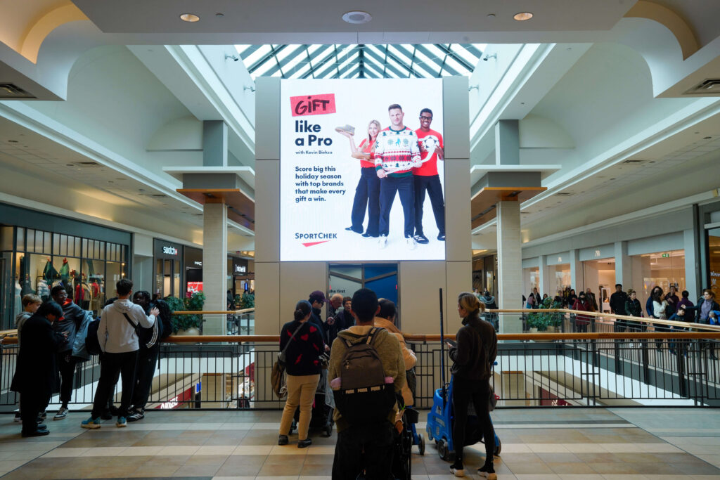 A busy two-level shopping mall atrium with shoppers gathered along the upper-floor railing. A large digital billboard dominates the center, displaying a SportChek holiday advertisement beneath a glass skylight.