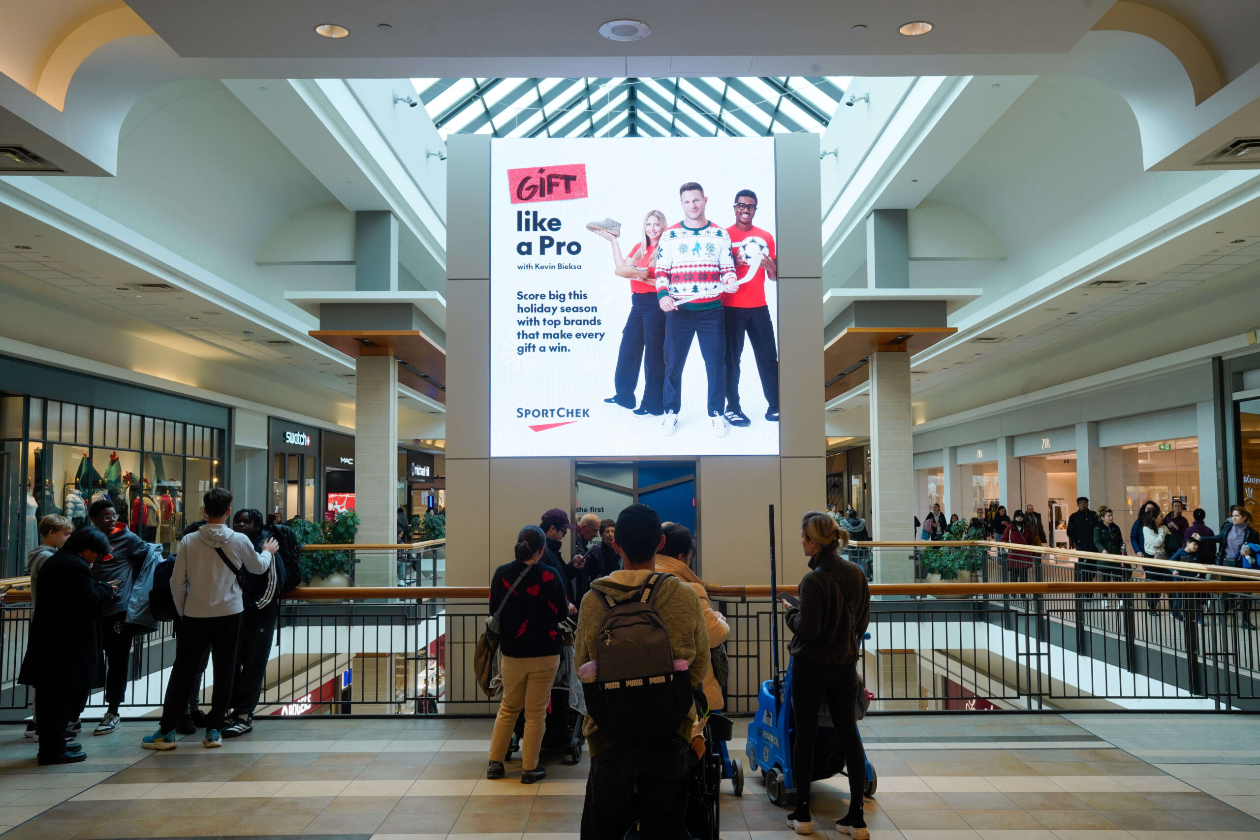 A busy two-level shopping mall atrium with shoppers gathered along the upper-floor railing. A large digital billboard dominates the center, displaying a SportChek holiday advertisement beneath a glass skylight.